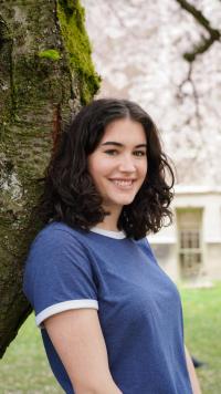 A young woman with curly hair smiles while leaning against a tree, surrounded by pink blossoms.